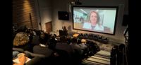 View from the rear of the audience of an event taking place on stage. An additional remote speaker is displayed on a large screen.