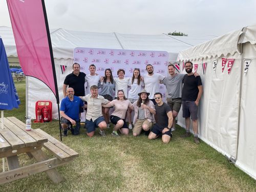 Thirteen people smile in amongst some gazebos, with their arms around each other's shoulders.