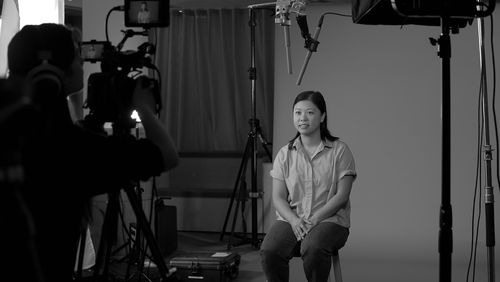 An interviewee sits on a stool in front of a plain backdrop. In the foreground, a camera operator adjusts their equipment.