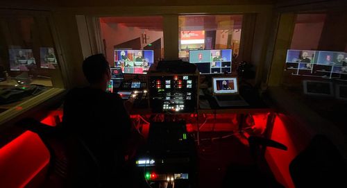 Photo from inside the technical booth for an event on stage. A person is operating the board, and the ongoing event is visible through a window.