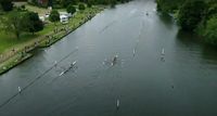 A drone shot of a river, with two rowing boats racing. A small crowd is gathered along the shore.