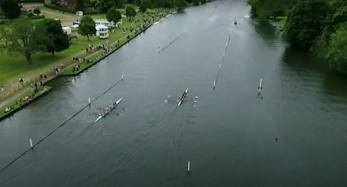 A drone shot of a river, with two rowing boats racing. A small crowd is gathered along the shore.