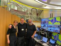 Three people with their arms around each other's shoulders stand in front of a technical event desk.