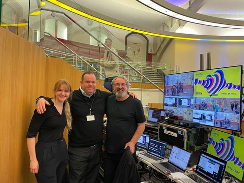 Three people with their arms around each other's shoulders stand in front of a technical event desk.