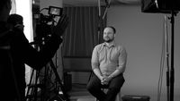 An interviewee sits on a stool in front of a plain backdrop. In the foreground, a camera operator adjusts their equipment.