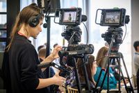 A camera operator stands behind two cameras. In the background an audience is visible.