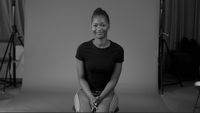 An interviewee sits on a stool in front of a plain backdrop.