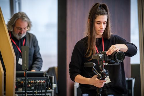 In the foreground, a camera operator adjusts a camera. In the background, a board operator adjusts a board.