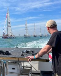 A person wearing a cap looks out to sea at a line of ships. A drone is on a table.