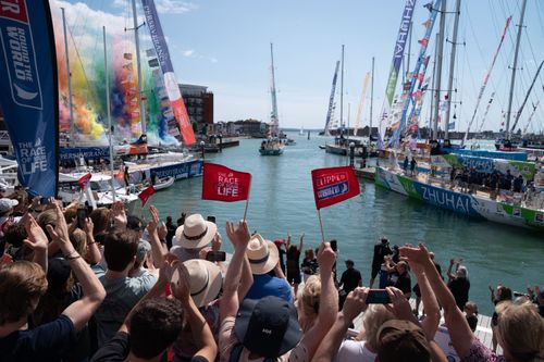 A celebrating crowd looks out at ships. A series of colourful towers of smoke have gone off to the left.