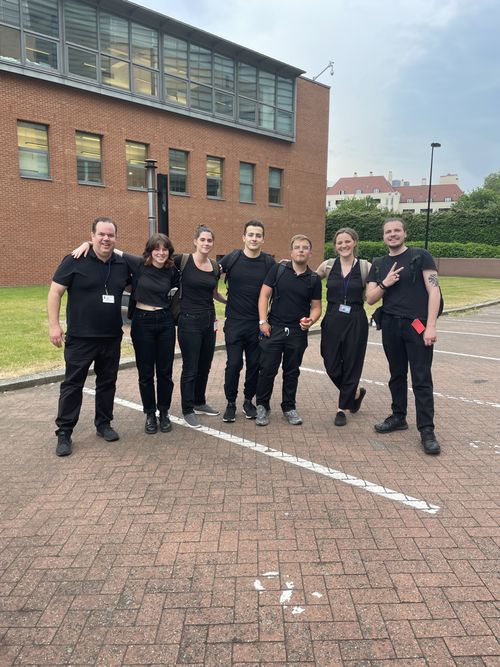 Seven people posing with their arms around each other's shoulders in a car park in front of a building.