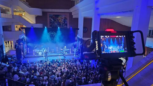 View from the rear of the crowd of a live musical performance. A monitor in the foreground shows the live event feed.