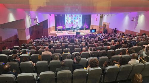 View from the rear of the room of an audience and stage. On stage, Joan Armatrading is being interviewed.